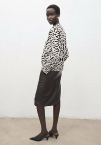 Sweter w zwierzęcy wzór w odcieniach beżu i czerni, zestawiony z dopasowaną, ciemną skórzaną spódnicą. Modelka ma na sobie czarne, szpiczaste buty na obcasie typu slingback. Gładkie faktury.