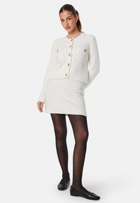 Vit tweed tvådelad outfit med en kavaj med guldknappar och en figursydd kjol. Modellen har svarta strumpbyxor och ballerinas, i stående pose.