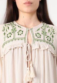 Blouse beige avec broderie florale verte, bords froncés et fermeture à nouer. Présente des à-côtés décoratifs et un tissu doux et texturé.