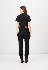 Maglietta nera e jeans blu scuro con cuciture visibili. I jeans presentano un'etichetta rossa "HUGO" sulla cintura posteriore. Tessuto liscio e vestibilità slim.