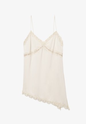 Femme aux longs cheveux foncés se tient contre un mur blanc, portant un pantalon ample couleur crème et un haut sans manches crème orné de dentelle.