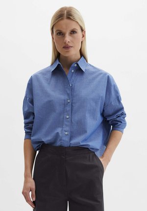 Blondynka w luźnej, niebieskiej koszuli zapinanej na guziki, włożonej do czarnych spodni z wysokim stanem, z jedną ręką w kieszeni.