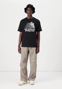 Svart t-skjorte med hvit grafikk og tekst, beige løstsittende bukser og kremhvite sneakers. Modellen har på seg en svart bøttehatt.