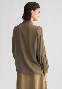 Blusa verde bronzo con colletto, maniche lunghe a palloncino e dettagli a bottoni sulle spalle. Tessuto morbido e fluido.