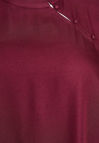 Burgundská blůza se hladkým povrchem, s polo zapínáním a mírně splývavým designem límce.