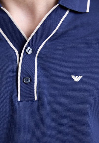 Tricou polo naval cu margini albe, având o deschidere cu două butoane și un logo brodat. Materialul are o textură netedă cu un guler tricotat plat.