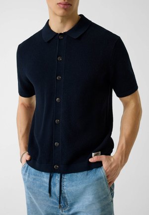 Homme portant un polo en maille à manches courtes noir avec col et boutons, et un jean bleu clair, les mains dans les poches.