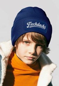 Navy knit beanie with a logo patch reading "Frechdachs." Worn over an orange turtleneck and a fleece-lined jacket. Close-up view.
