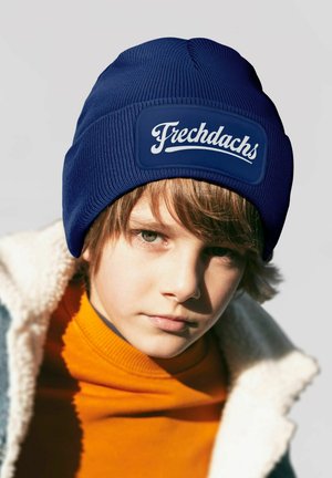 Marineblaue Strickmütze mit einem Logo-Patch, das "Frechdachs" zeigt. Getragen über einem orangefarbenen Rollkragenpullover und einer mit Fleece gefütterten Jacke. Nahaufnahme.
