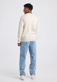 Light beige sweatshirt with ribbed cuffs and hem, paired with light blue, loose-fitting denim jeans and white sneakers. Plain background.