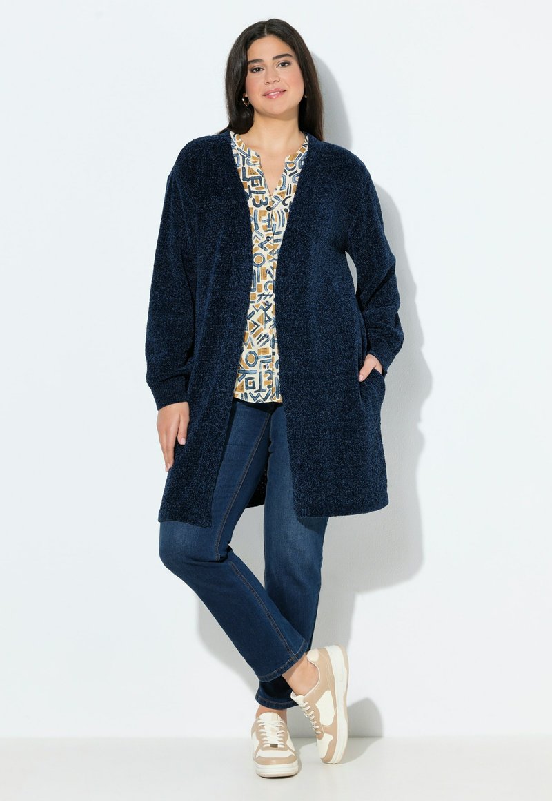 Tummansininen tekstuurinen pitkä kardigan, jossa on avonaiset etuosat, kaksi taskua, käytetty kuviollisen paidan päällä, yhdistetty tummiin farkkuihin ja vaaleanbeigeihin lenkkareihin.