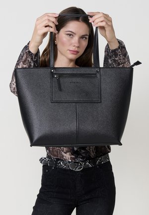 Mujer sosteniendo un gran bolso de cuero negro con un bolsillo frontal con cremallera, vistiendo una blusa de estampado de serpiente, un cinturón a juego y pantalones negros.