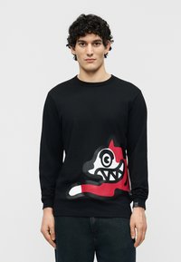 Νεαρός με σγουρά μαλλιά, φορώντας μαύρο μακρυμάνικο πουκάμισο με κόκκινο, λευκό και μαύρο γραφικό μιας στιλιζαρισμένης ζωγραφιάς ζώου.