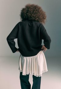 Persona con capelli ricci di spalle, che indossa un maglione nero sopra una gonna bianca con pizzo e pantaloni scuri, con le mani appoggiate sui fianchi.