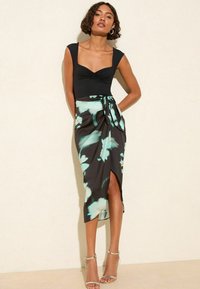 Haut noir ajusté avec décolleté carré, associé à une jupe portefeuille à imprimé floral dans des tons sombres et des accents bleu clair. Sandales en argent à lanières.