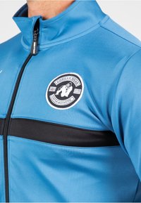 Blauwe sportjack met opstaande kraag, volledige rits, zwarte horizontale streep en een ronde logo met een gorilla-ontwerp. Glad van textuur.