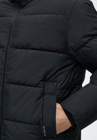 Veste matelassée noire avec fermeture éclair frontale, coutures visibles, petit logo sur la poitrine et main insérée dans la poche latérale.