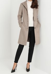 Beige wool blend coat with a lapel collar, two front pockets, and a single button closure, worn over a light sweater and black trousers.