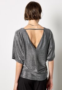 Glänzendes silbernes Oberteil mit kurzen Ärmeln, tiefem V-Ausschnitt hinten und gerafftem Bund. Hergestellt aus einem metallischen Stoff mit glatter Textur. Einfaches und elegantes Design.