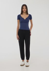 Top corto aderente blu navy con scollatura a V e dettagli di bottoni, abbinato a pantaloni in stile jogger neri e sneakers bianche con suola marrone.