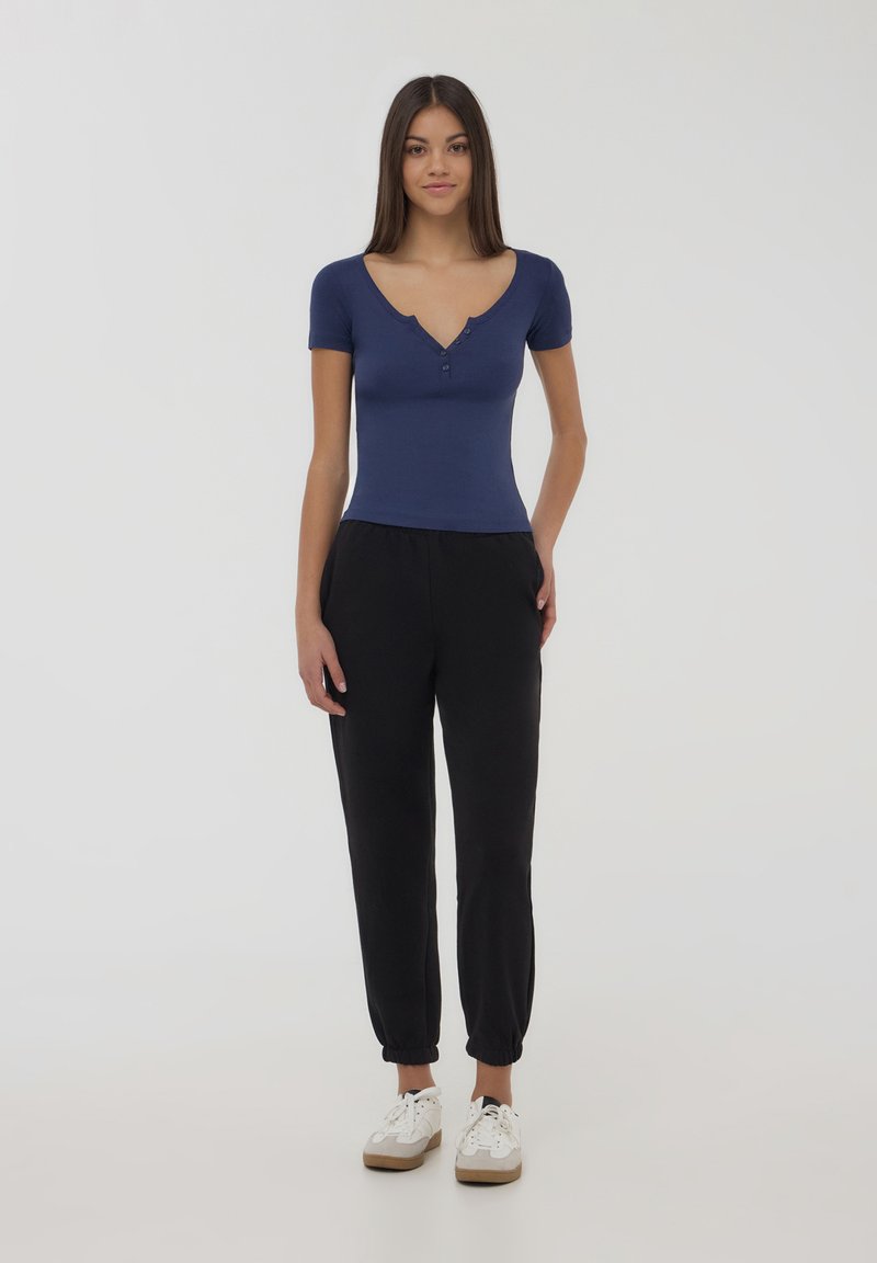 Top corto aderente blu navy con scollatura a V e dettagli di bottoni, abbinato a pantaloni in stile jogger neri e sneakers bianche con suola marrone.
