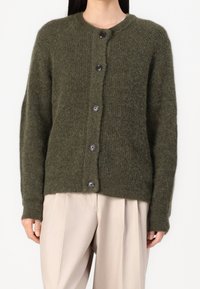 Olivewoska zielona kardiganka wykonana z teksturowanej dzianiny, z okrągłym dekoltem, guzikami z przodu i długimi rękawami, w zestawie z beżowymi spodniami.