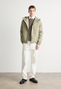 Light olive green jacket with a hood, zip closure, and elastic cuffs, worn over a brown sweater and white trousers; black shoes complete the outfit.