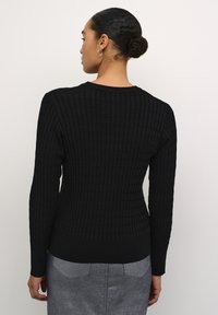Maglione nero a coste con maniche lunghe, scollatura tonda e motivo testurizzato. Silhouette aderente che si restringe in vita.