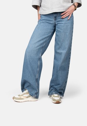 Persona que lleva vaqueros anchos de color azul claro, zapatillas beige y blancas, y una sudadera gris con las manos en los bolsillos.