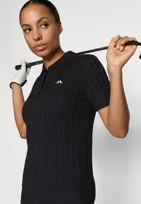 Polo de golf de punto negro con un patrón de trenzas, mangas cortas y un dobladillo acanalado. Presenta un pequeño logo blanco en el collar.