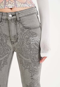 Jeans in denim grigio con intricate ricami neri, vita alta, cinque tasche e chiusura frontale con bottone.