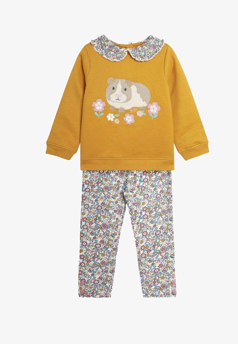 Keltainen huppari, jossa on marsuvanhustyylinen kuvio, kukka-akcentteja ja röyhelökaulus, yhdistettynä valkoisiin housuihin, joissa on monivärinen kukkakuosi.