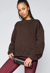 Sweatshirt oversized castanho com punhos e bainha canelados, usado com leggings bordeaux. Uma garrafa de água preta é segurada na mão esquerda da modelo.