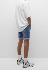 Valkoinen ylisuuressa lyhythihaisessa t-paidassa, farkkushortseissa, joiden lahkeet on käännetty, ja urheilukengissä, joissa on monivärinen kuviointi ja valkoiset sukat.