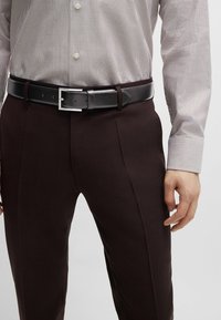 Black leather belt with a silver rectangular buckle, worn over dark burgundy textured trousers and a light striped button-up shirt.