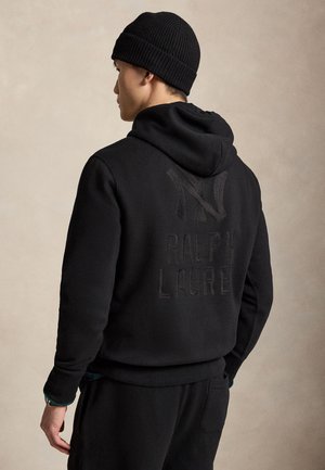 Jeune homme portant un bonnet noir et un sweat noir avec le texte et le logo Ralph Lauren dans le dos, vu de dos sur un fond uni.