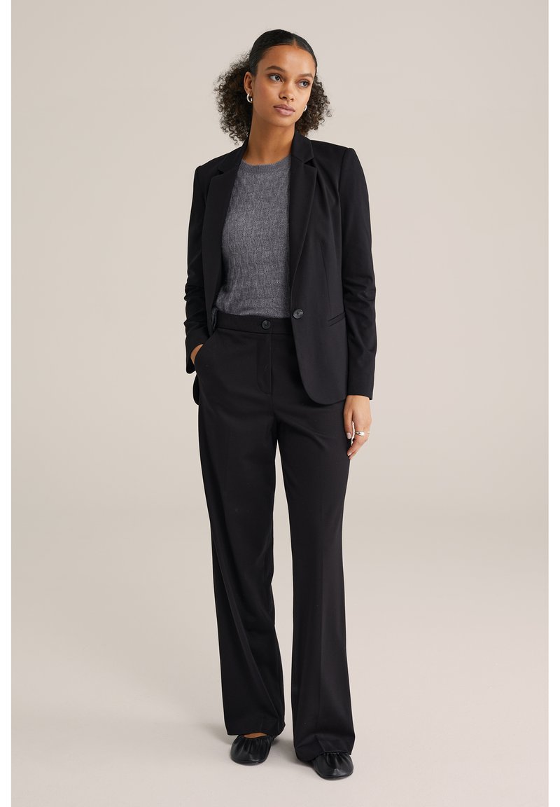 Čierny priliehavý oblek pozostávajúci z prispôsobeného saku a nohavíc s širokými nohavicami; doplnený o štruktúrovaný sivý sveter a čierne ploché topánky.