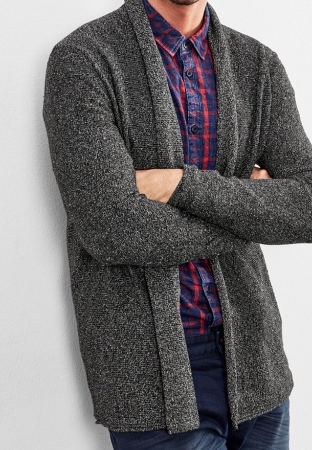 Homme les bras croisés portant un cardigan gris foncé ouvert sur une chemise à carreaux rouge et bleue boutonnée et un pantalon foncé.