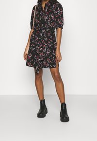 Černé květinové šaty s krátkými rukávy, zdobené vícolorými květinovými vzory, s zavazováním v pase a zapínáním na knoflíky, doplněné černými kotníkovými botami.