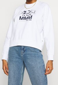 Vit sweatshirt med tryckt grafik av Disney-karaktärer och texten "MMM", med avslappnad passform och ribbade ärmslut. Matchad med blå jeans.
