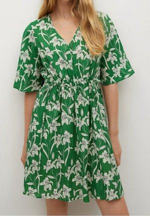 Femme portant une robe verte à manches courtes avec un imprimé floral blanc et une taille cintrée, debout contre un fond uni.