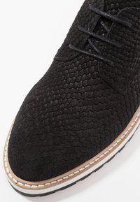 Scarpa nera in suede con una superficie testurizzata e squamosa, design con lacci e una suola cucita a contrasto in tan e bianco. Punta rotonda.