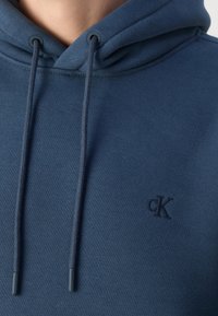 Personne portant un sweat à capuche bleu avec cordons et un petit logo '"cK"' brodé sur la poitrine.