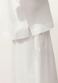 Gros plan sur un haut en lin blanc avec une fente latérale porté avec une jupe en lin assortie taille haute, sur un fond uni.