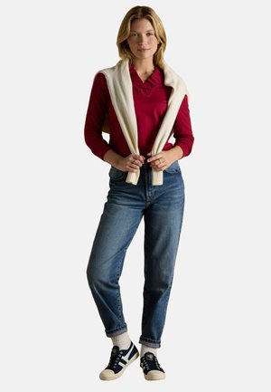 Red long-sleeve top with ruffled collar, cream sweater draped over shoulders, blue high-waisted jeans, and navy sneakers with cream soles.