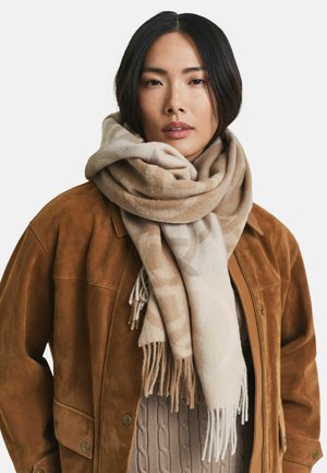 Jeune femme portant un grand foulard en beige à motifs, une veste en daim marron et un pull en tricot torsadé taupe sur fond blanc.