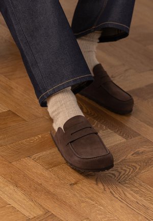 Mocassini in camoscio marrone indossati con calzini beige a coste e jeans di denim blu scuro su un pavimento in parquet di legno.