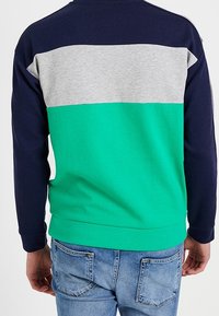 Mies, joka käyttää sinivihreä-harmaa väreissä väriblocki collegepaitaa, sinisten farkkujen kanssa, näkyy selkäpuolelta yksinkertaista taustaa vasten.