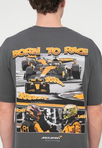 Γκρι μπλούζα με πολύχρωμο γραφικό εκτύπωσης αγωνιστικών αυτοκινήτων και δύο οδηγούς με κράνη, με το κείμενο "ΓΕΝΝΗΜΕΝΟΣ ΓΙΑ ΝΑ ΤΡΕΧΕΙ" και "SKRRRRT."