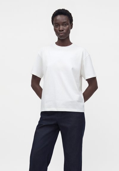 Woman standing with hands behind back wearing a plain white short-sleeve shirt and dark navy pants against a plain light background.